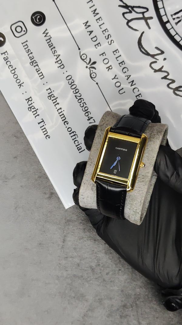 A rectangular watch with a gold-tone case, black dial, and black leather strap, displayed on a gray cushion held by a gloved hand. A branded paper card is visible in the background.